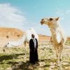 Jessica Nabongo with two camels in the desert, wearing a long black robe and a black and white scarf draped around her head