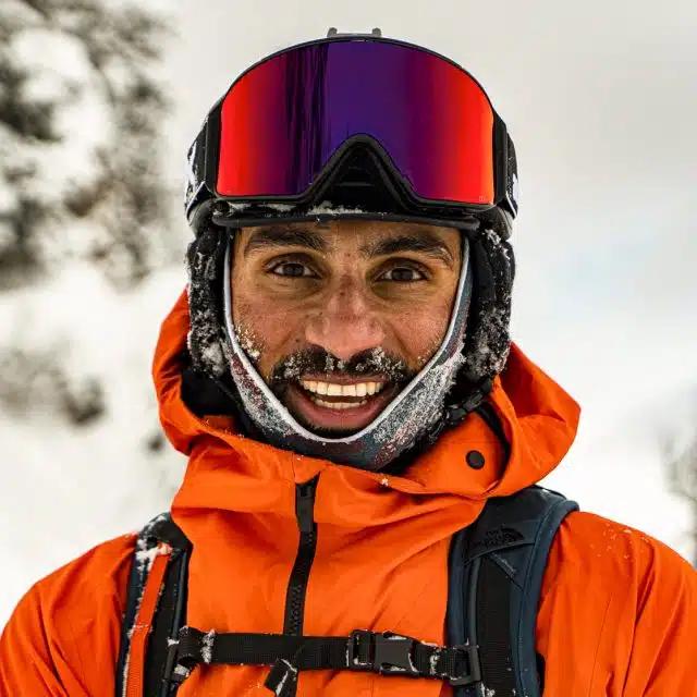 Pro skier and accessibility activist Vasu Sojitra wearing reflective ski goggles and an orange ski jacket