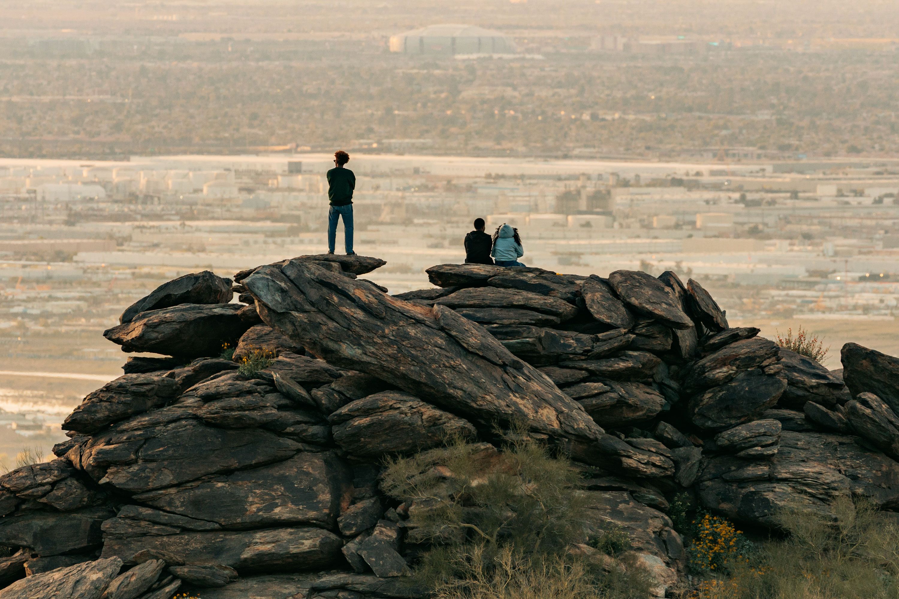 People perched on large desert boulders looking out over an expansive urban landscape in hazy evening light.