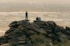 People perched on large desert boulders looking out over an expansive urban landscape in hazy evening light.