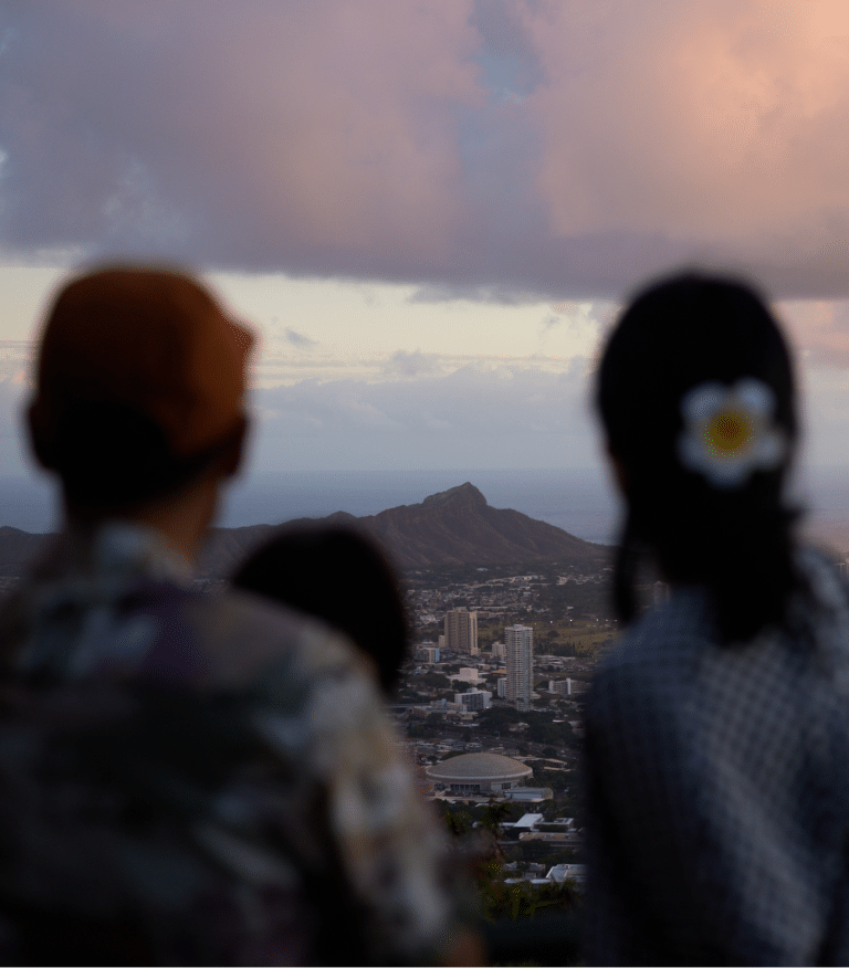 A couple, one with a flower in her hair, look over the skyline of Honolulu to mountains beyond