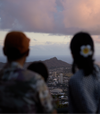 A couple, one with a flower in her hair, look over the skyline of Honolulu to mountains beyond