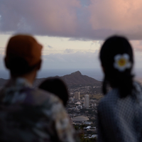 A couple, one with a flower in her hair, look over the skyline of Honolulu to mountains beyond