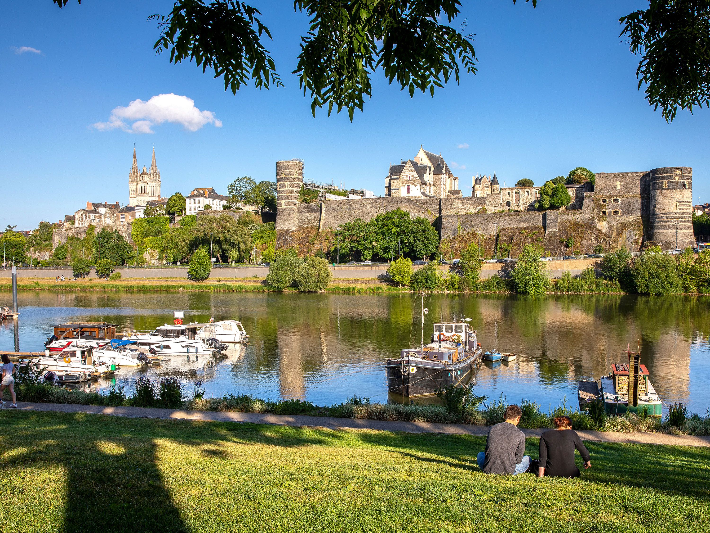 Two people sit along the water with boats docked and medieval buildings.
