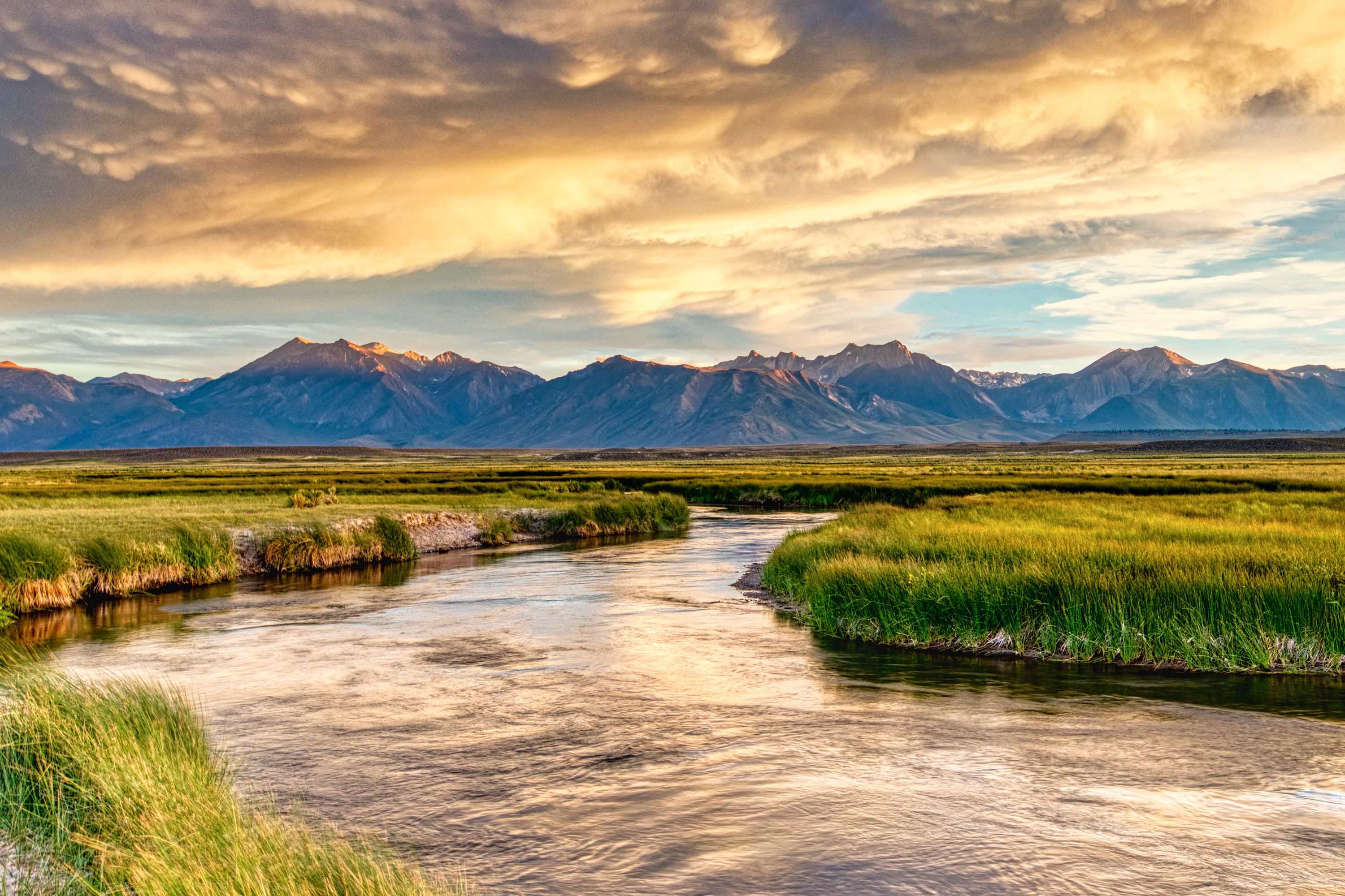 A winding river flows through grassy plains beneath dramatic clouds and distant mountain peaks.