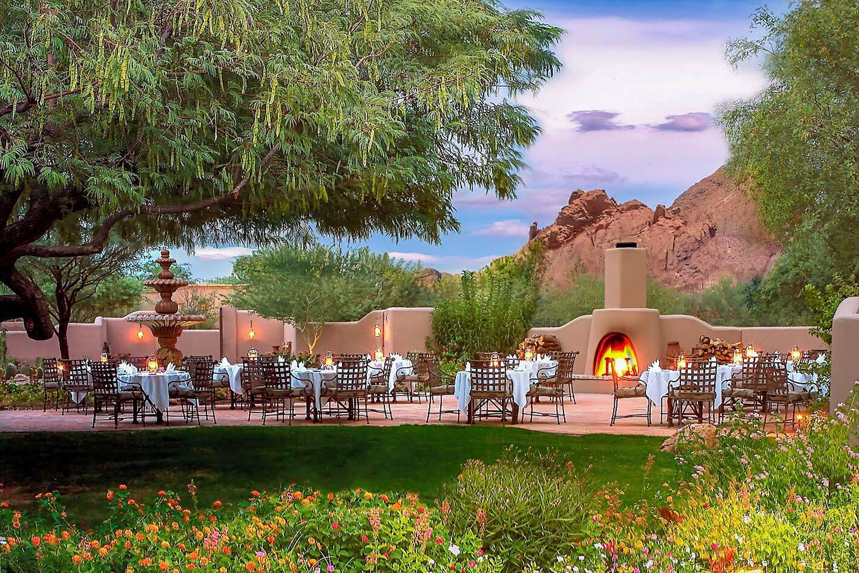 A brick walkway in the foreground gives way to wild flowers, and an outdoor patio dining area with an adobe fireplace.