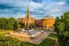 Aerial view of memorial structure in park with city skyline in the background under cloudy sky.