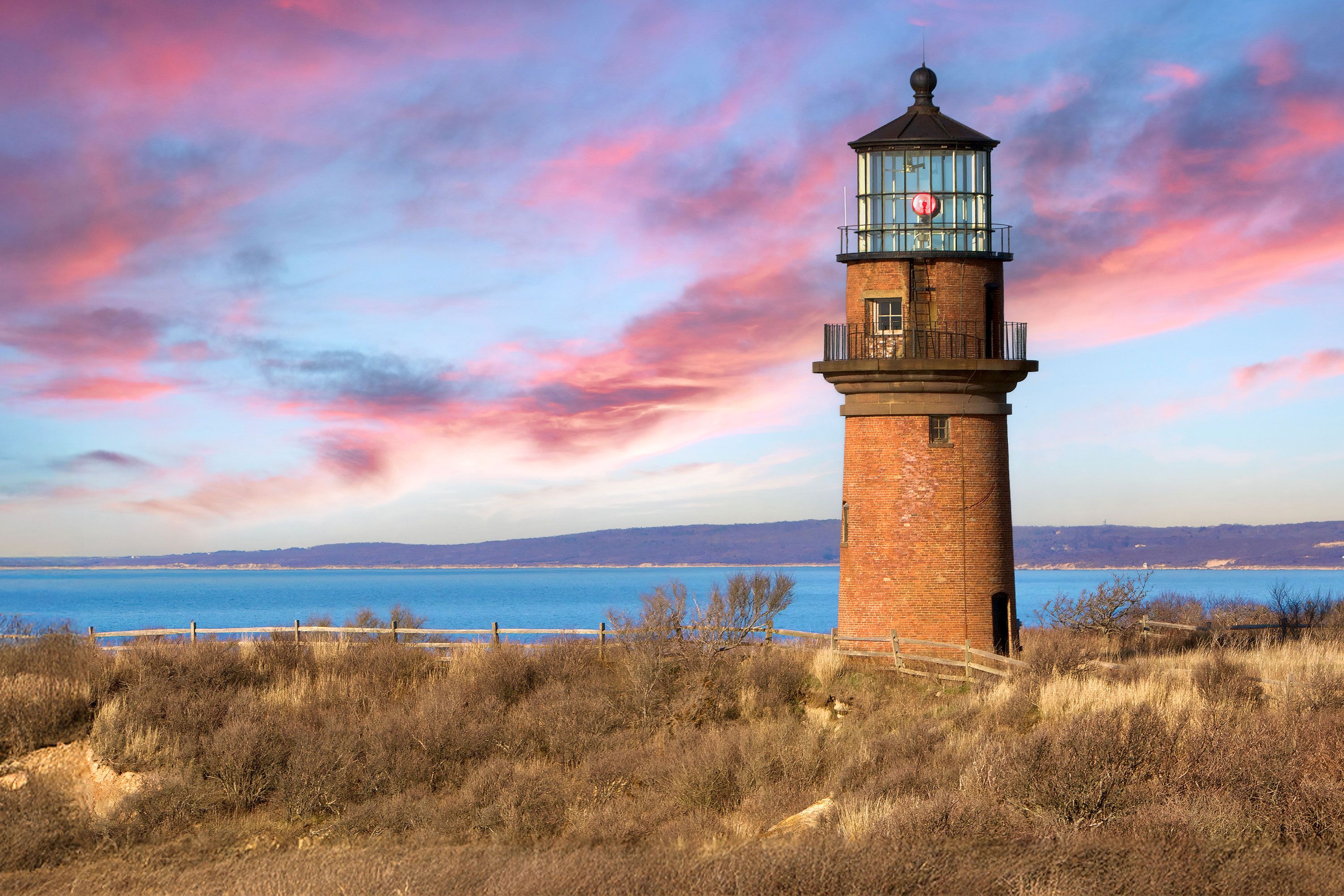 A brick lighthouse standing on cliff-top above Martha’s Vineyard shoreline, bathed in warm orange and pink sunset light, with a glowing lantern room against a dusky sky over the ocean.