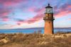 A brick lighthouse standing on cliff-top above Martha’s Vineyard shoreline, bathed in warm orange and pink sunset light, with a glowing lantern room against a dusky sky over the ocean.
