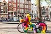 An old, weathered bicycle parked on a cobblestone street in Amsterdam, decorated with vibrant rainbow streamers, with historic European-style buildings blurred in the background.
