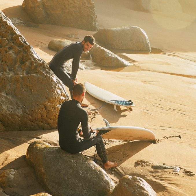 Two surfers in wetsuits smile, sitting on big rocks, on the beach in Lisbon