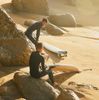 Two surfers in wetsuits smile, sitting on big rocks, on the beach in Lisbon