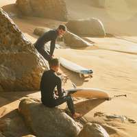 Two surfers in wetsuits smile, sitting on big rocks, on the beach in Lisbon