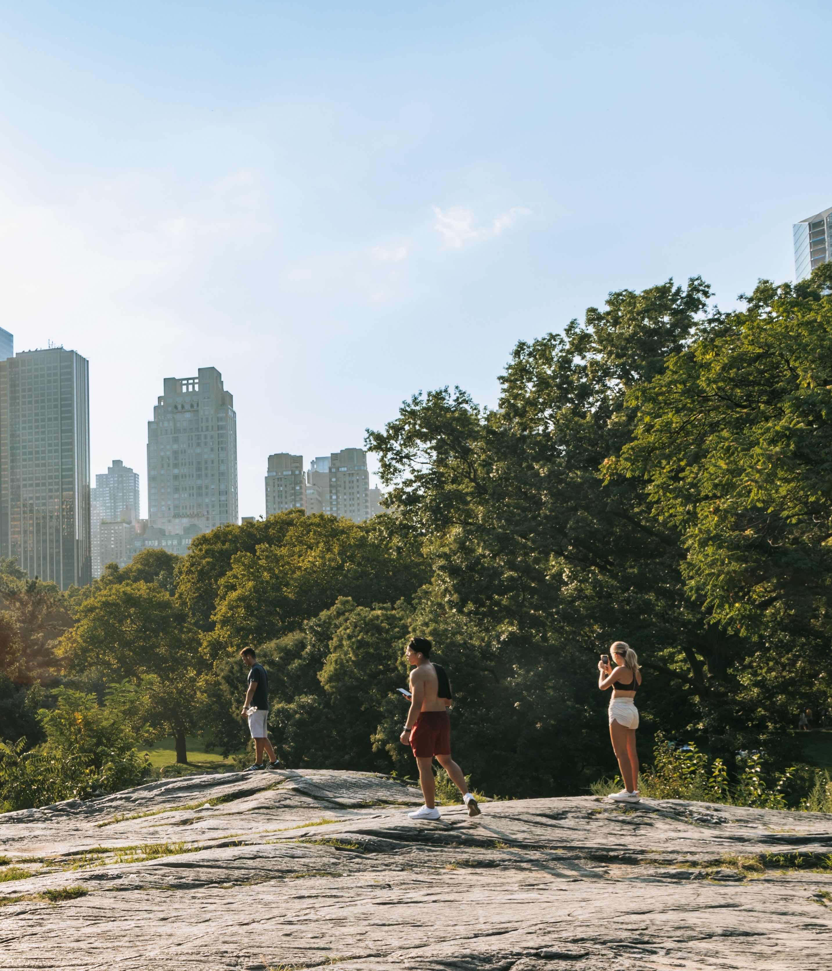 People stand atop the rocks in Central Park with the Manhattan skyline in the distance in New York City