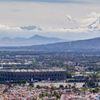 A wide shot of Mexico City shows houses to the foreground, the Azteca Stadium in the middle and cloud covered mountains in the background