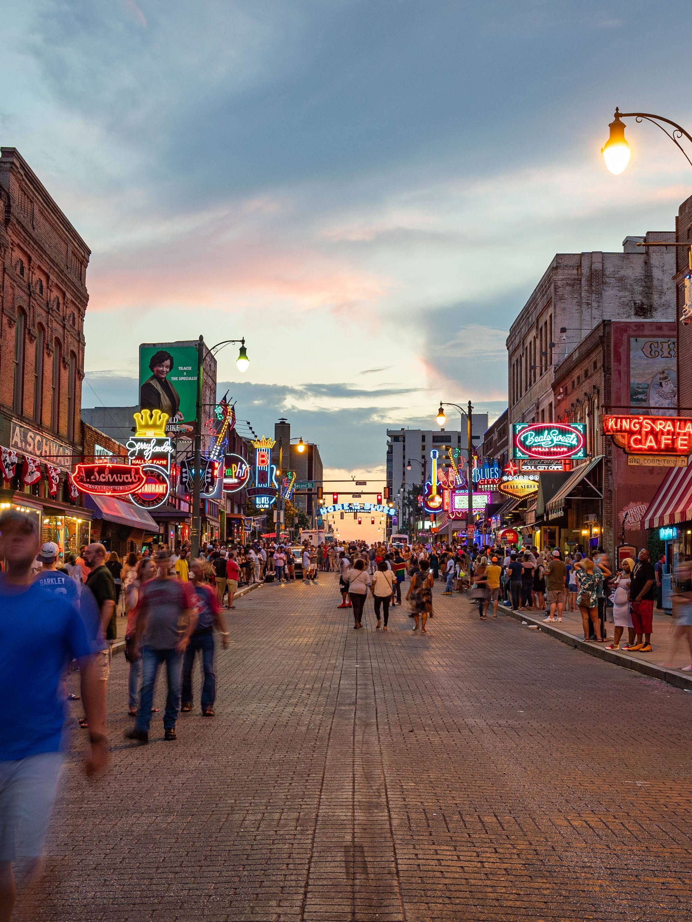 A busy street in Mississippi at dusk, with pedestrians walking past restaurants and shops with bright neon lights