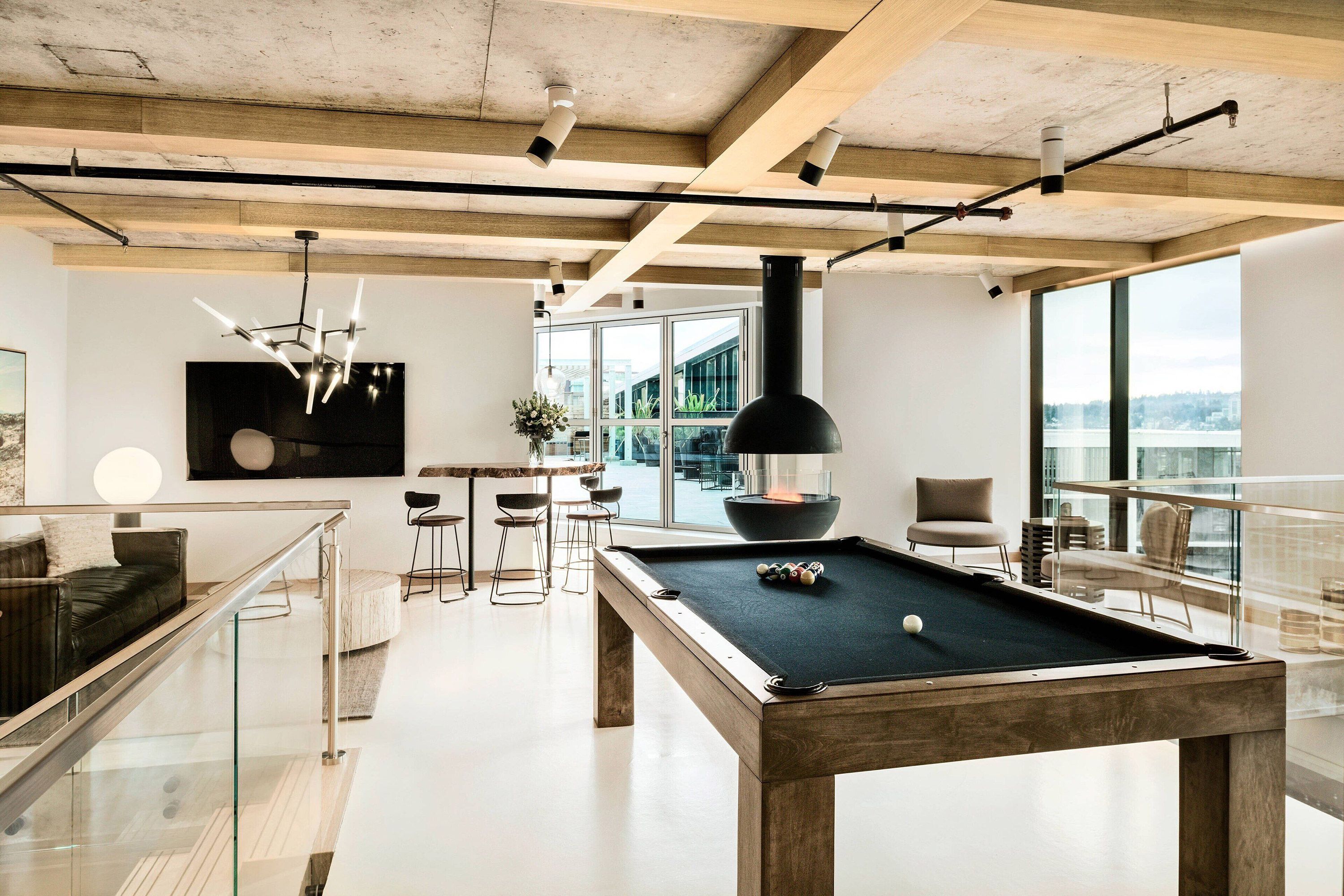 A pool table with a glass railing to the side, a modern table and chairs behind with a circular fireplace and wood ceiling beams.