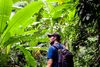 Man with backback and cap hikes through thick jungle.