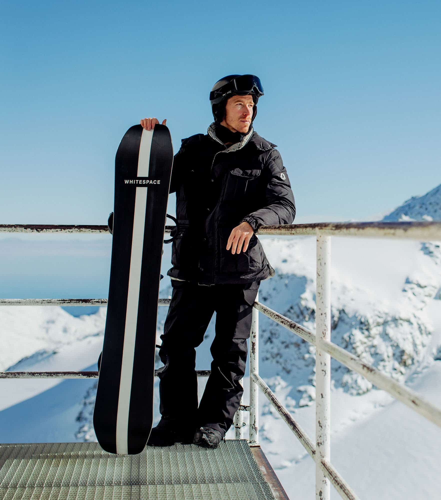 Shaun White holds his snowboard and leans against the railings of a platform surrounded by snowy mountains holds his snowboard