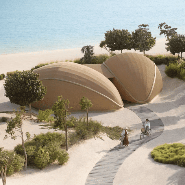 Two women cycle down a curved path away from two pod shaped brown buildings on the shore of the Red Sea