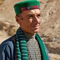 A man wearing a bright green and red embroidered hat and striped scarf squints into the sunlight