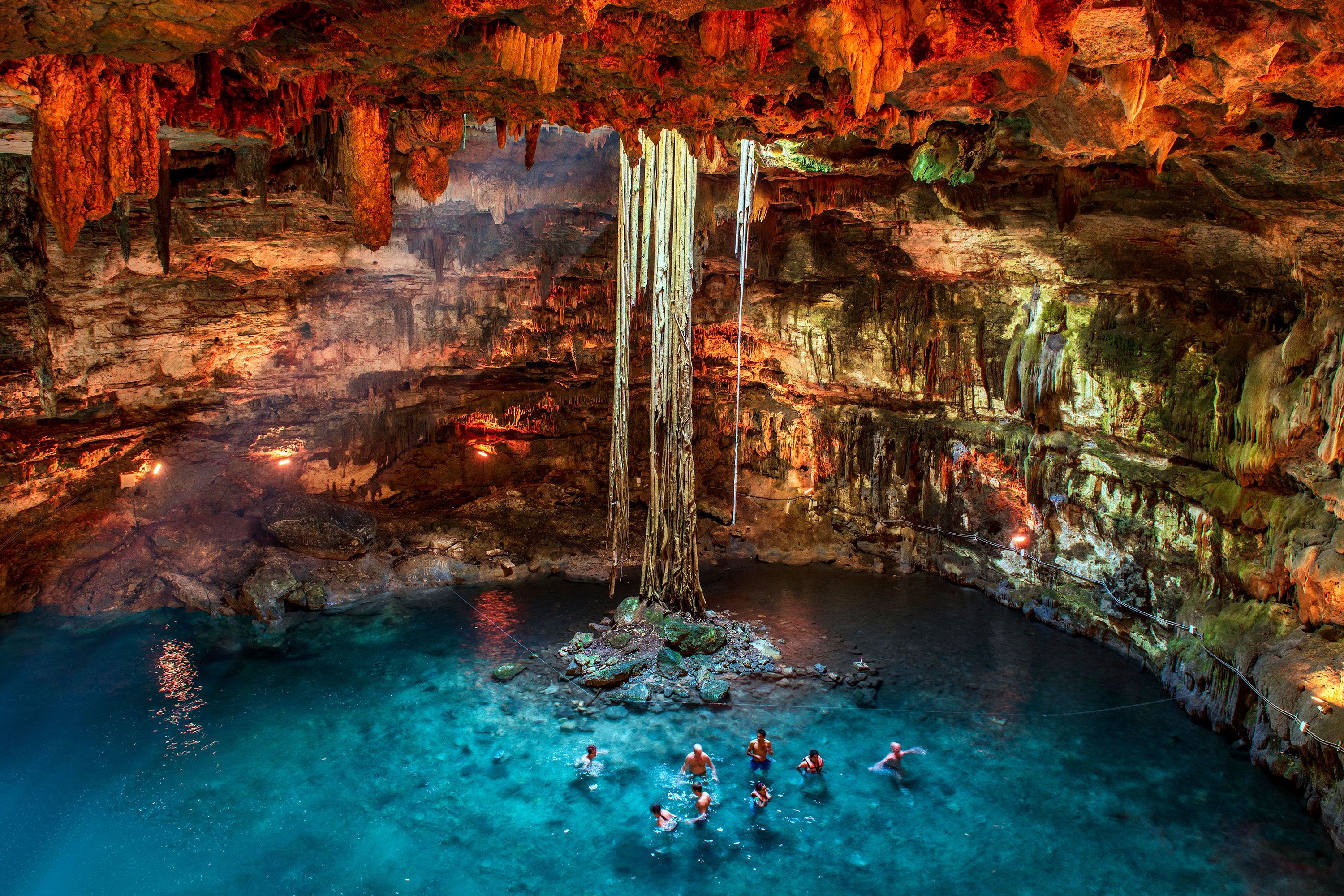 Swimmers wade in a glowing underground cenote beneath stalactites and hanging roots.