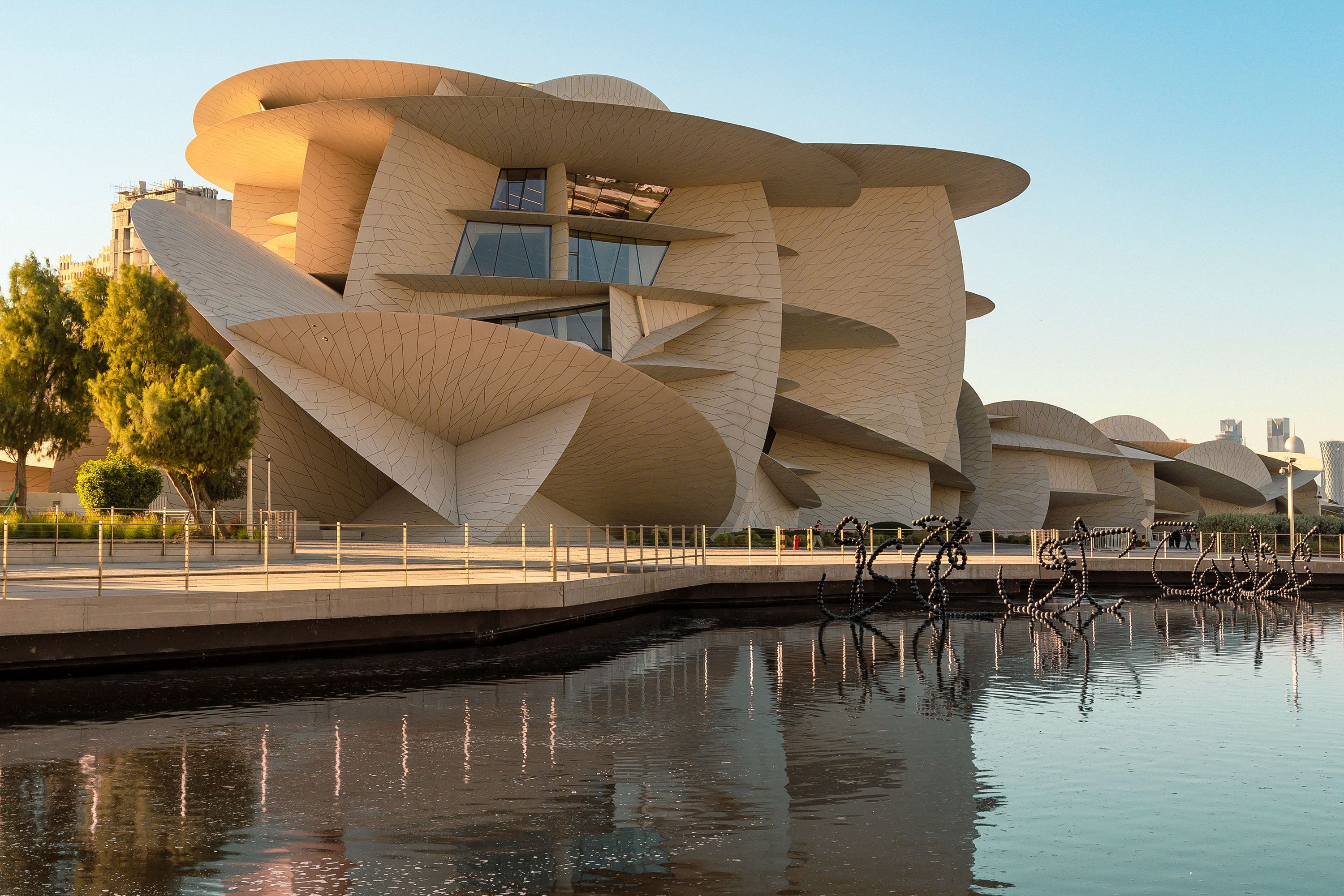A wide-angle view of the National Museum of Qatar’s sand-colored interlocking disc façade, with a still reflecting pool and greenery in the foreground, and Doha’s waterfront beyond.