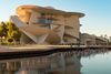 A wide-angle view of the National Museum of Qatar’s sand-colored interlocking disc façade, with a still reflecting pool and greenery in the foreground, and Doha’s waterfront beyond.