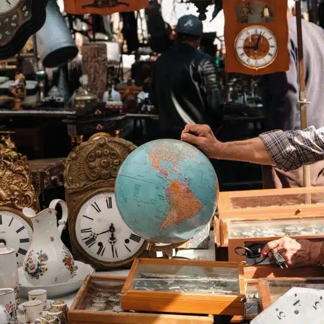 A man holds the top of a globe surrounding by clocks and other antiques at a flea market