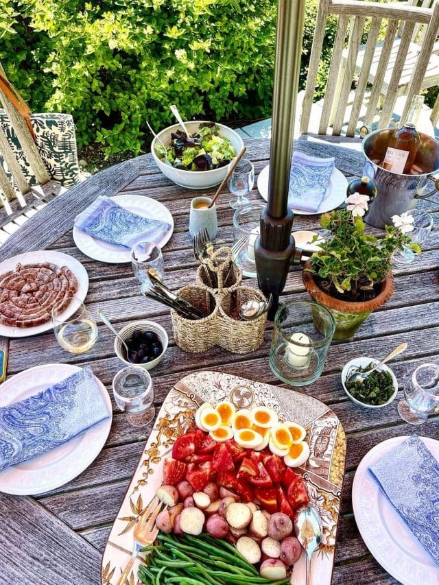 An al fresco lunch on a wooden table, set with pink plats and patterned blue napkins, condiments and a platter of boiled eggs and colorful vegetables