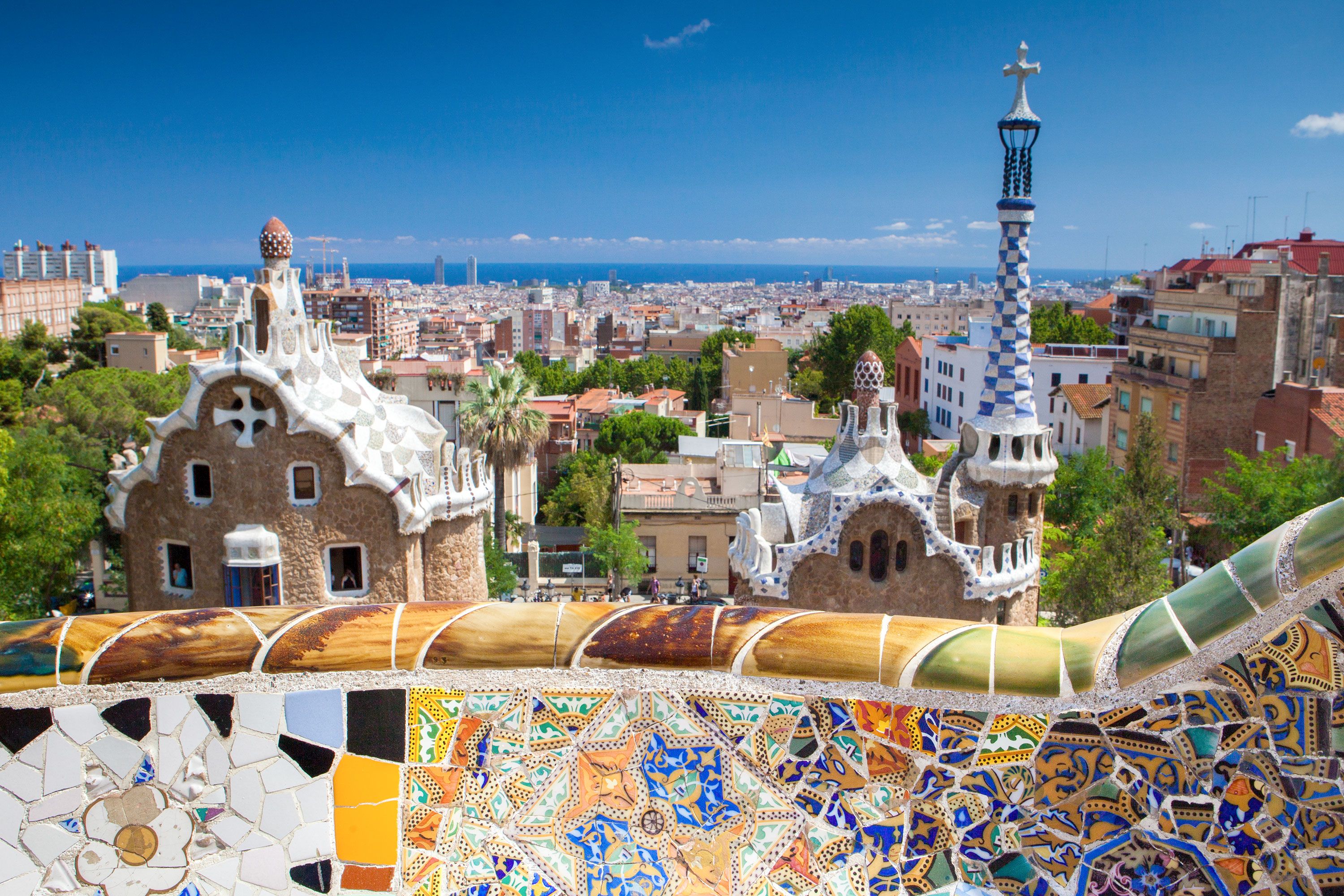 Colorful mosaic railing and Gaudí-designed towers at Park Güell overlooking Barcelona with the sea in the distance.