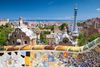 Colorful mosaic railing and Gaudí-designed towers at Park Güell overlooking Barcelona with the sea in the distance.