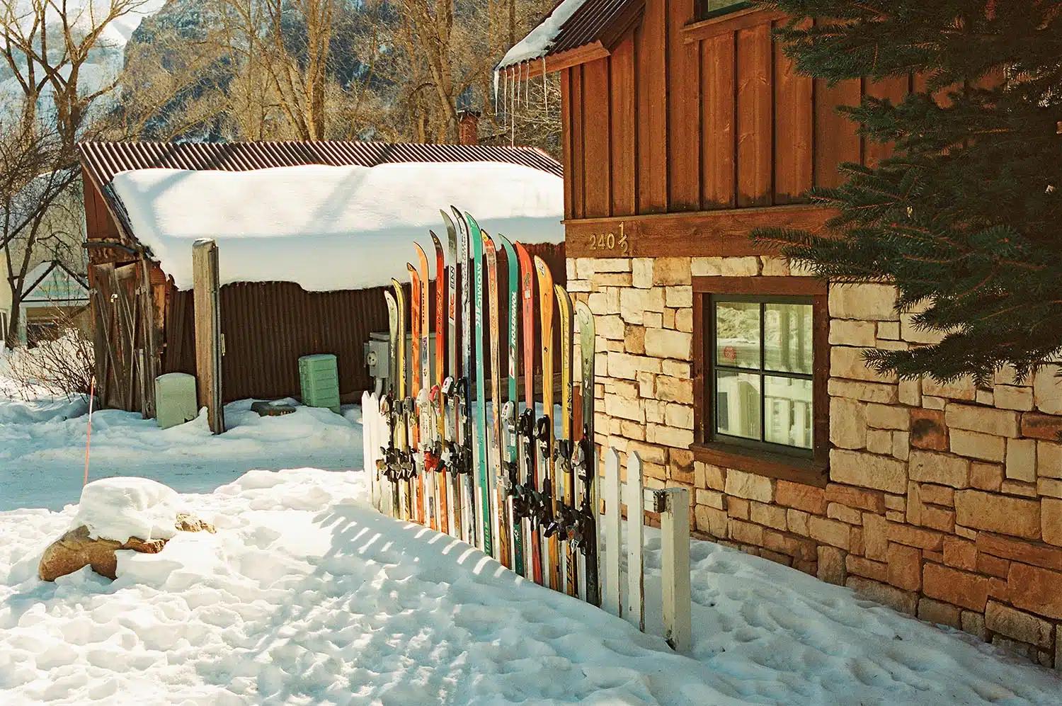 Colourful skis are held upright against a fence outside a chalet at a ski resort covered by thick snow