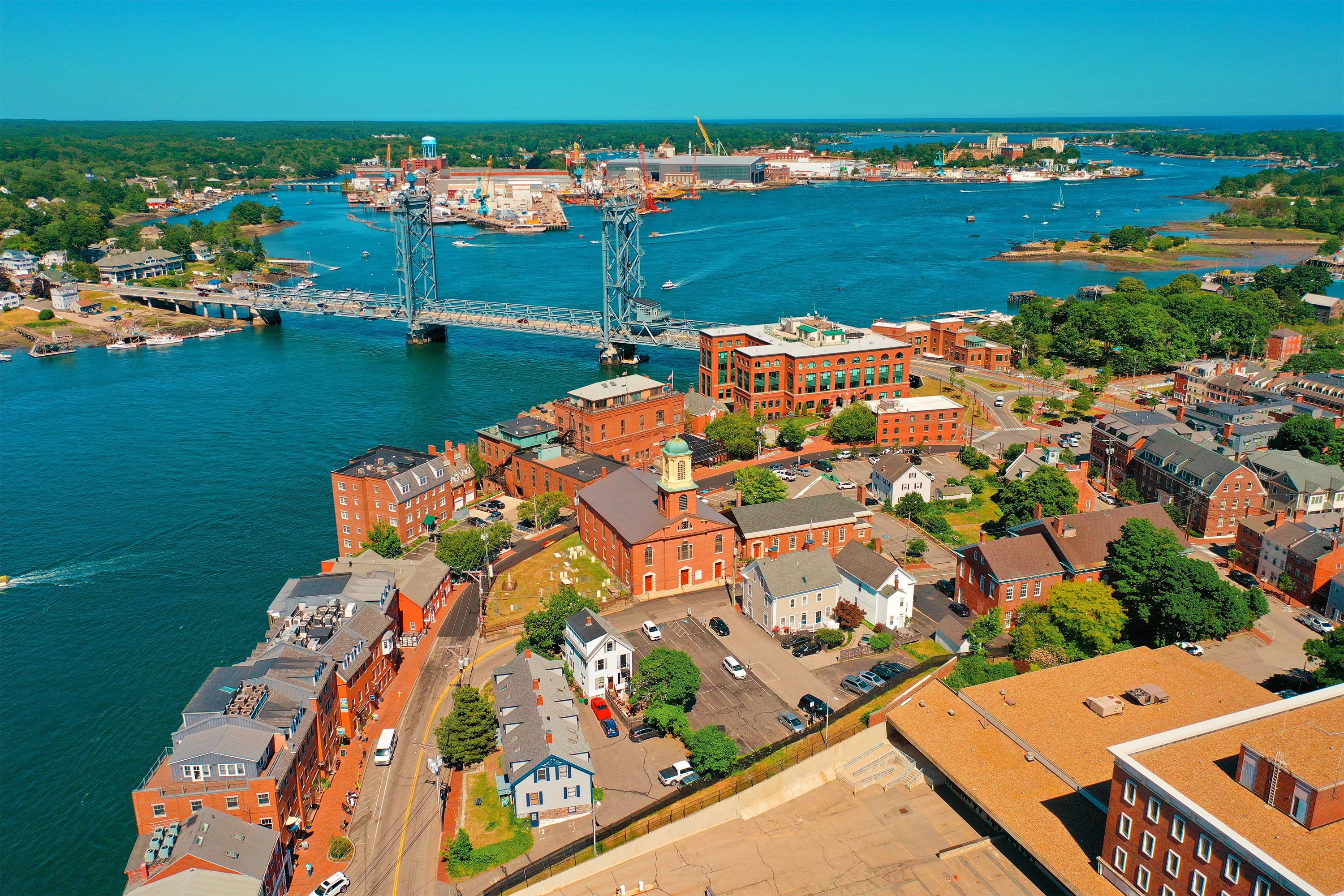 A drone view over downtown Portsmouth, NH, showing dense clusters of brick and wooden buildings, narrow streets, and a waterfront area, with a clear summer sky overhead.