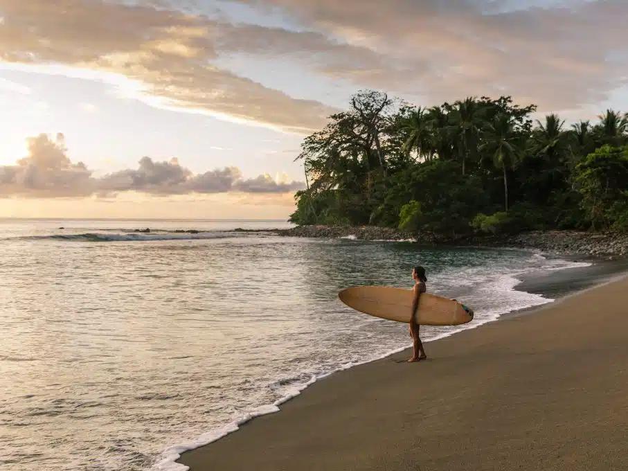 A woman holding a surf board under her left arm stands at the edge of the water looking out to the sea on a beach in Guanacaste, Costa Rica