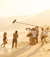 A man and woman in bathing suits emerge from the sea as a film crew stand in front of them filming