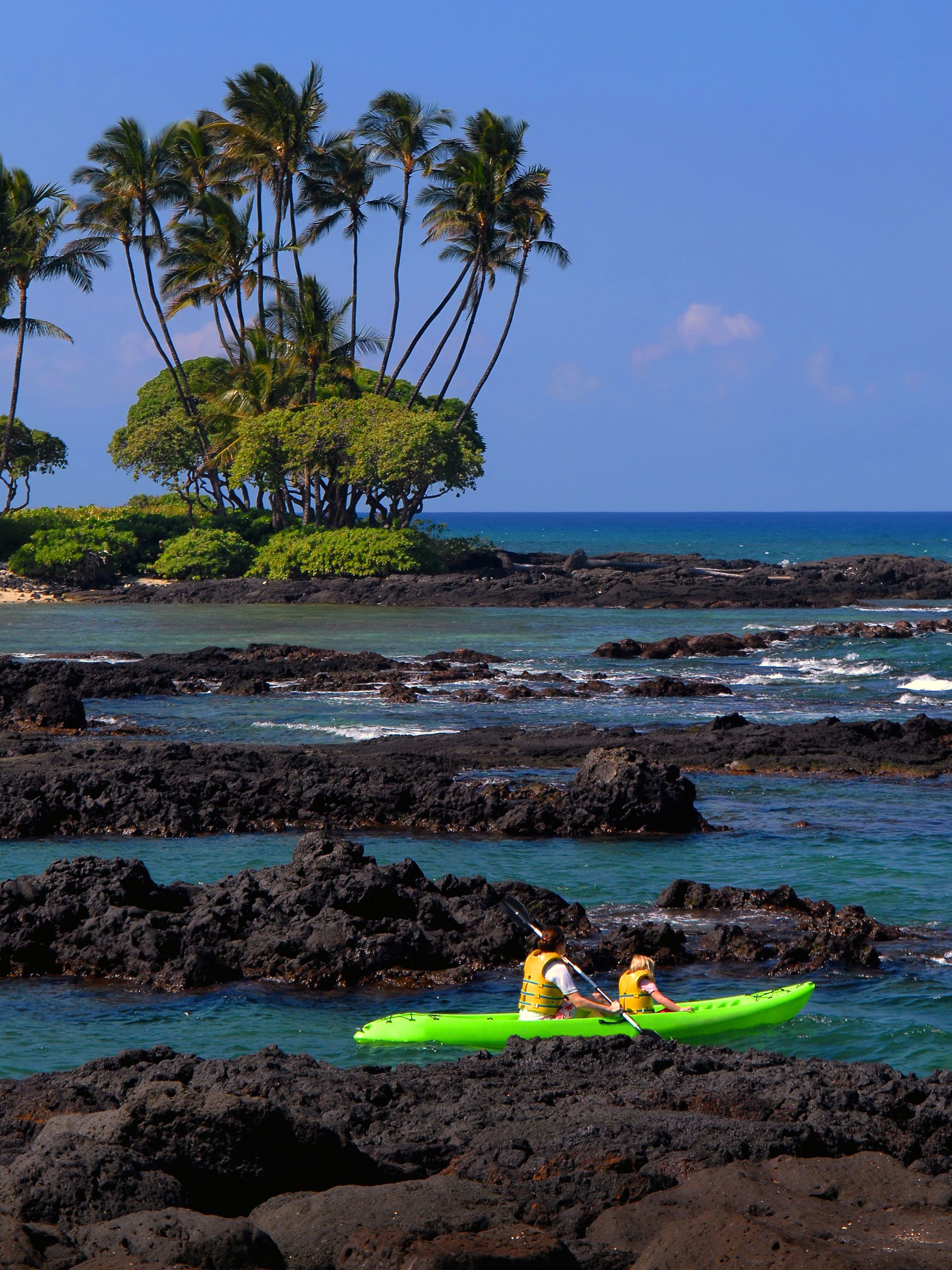 Two kayakers paddle past black lava rocks and palms in a calm tropical bay.