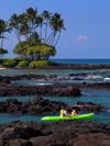 Two kayakers paddle past black lava rocks and palms in a calm tropical bay.