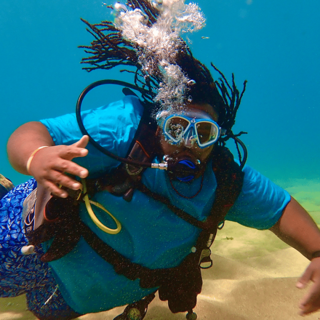 Travel influencer Jeff Jenkins wears diving gear, blue swimming trunks and a t-shirt as he swims underwater above the sandy ocean floor