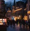 Shoppers walk a cobbled lane in [town], past stone houses and market chalets strung with Christmas lights at dusk.
