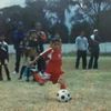 A slightly blurred and creased image of Edson Álvarez playing football as a child, in a red kit, kicking the ball with his right foot
