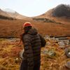 A person in a black puffer jacket and orange beanie hikes in the Scottish Highlands during fall