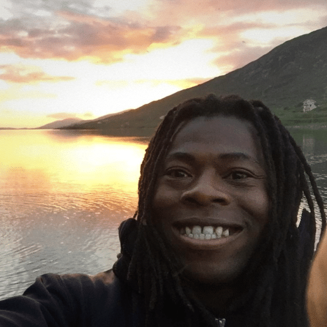 Ade Adepitan smiles for a selfie, a mountain and sea behind him