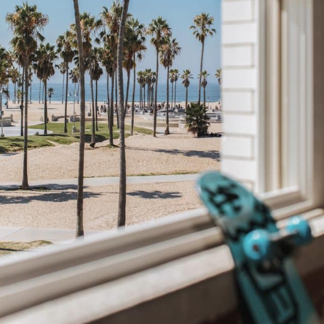 A view of the beach, palm trees, and the Pacific beyond, from the window of the Venice V Hotel; a skateboard leans against the windowsill