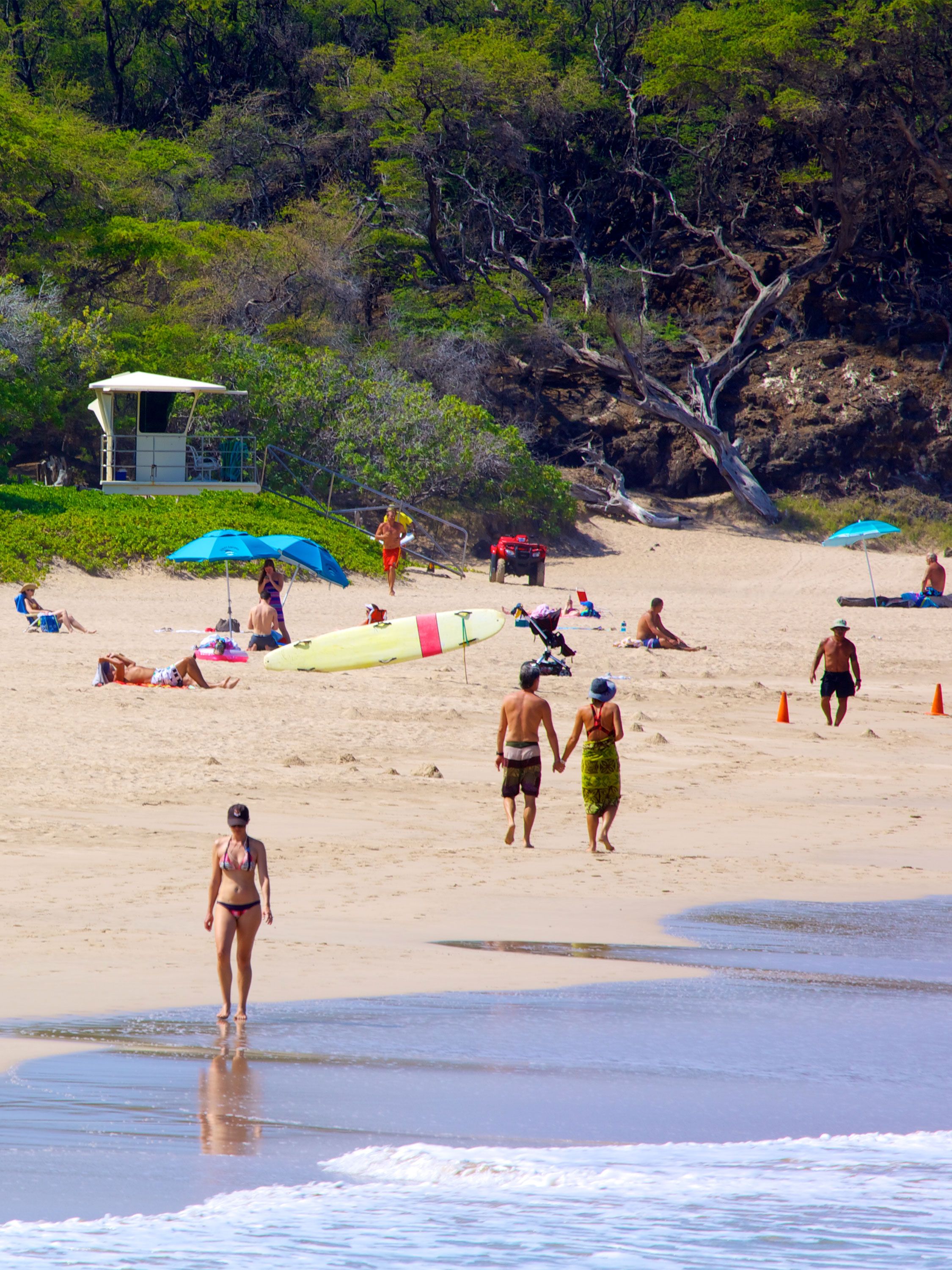 Beachgoers stroll and relax on a sunny Hawaiian shoreline backed by lush cliffs.