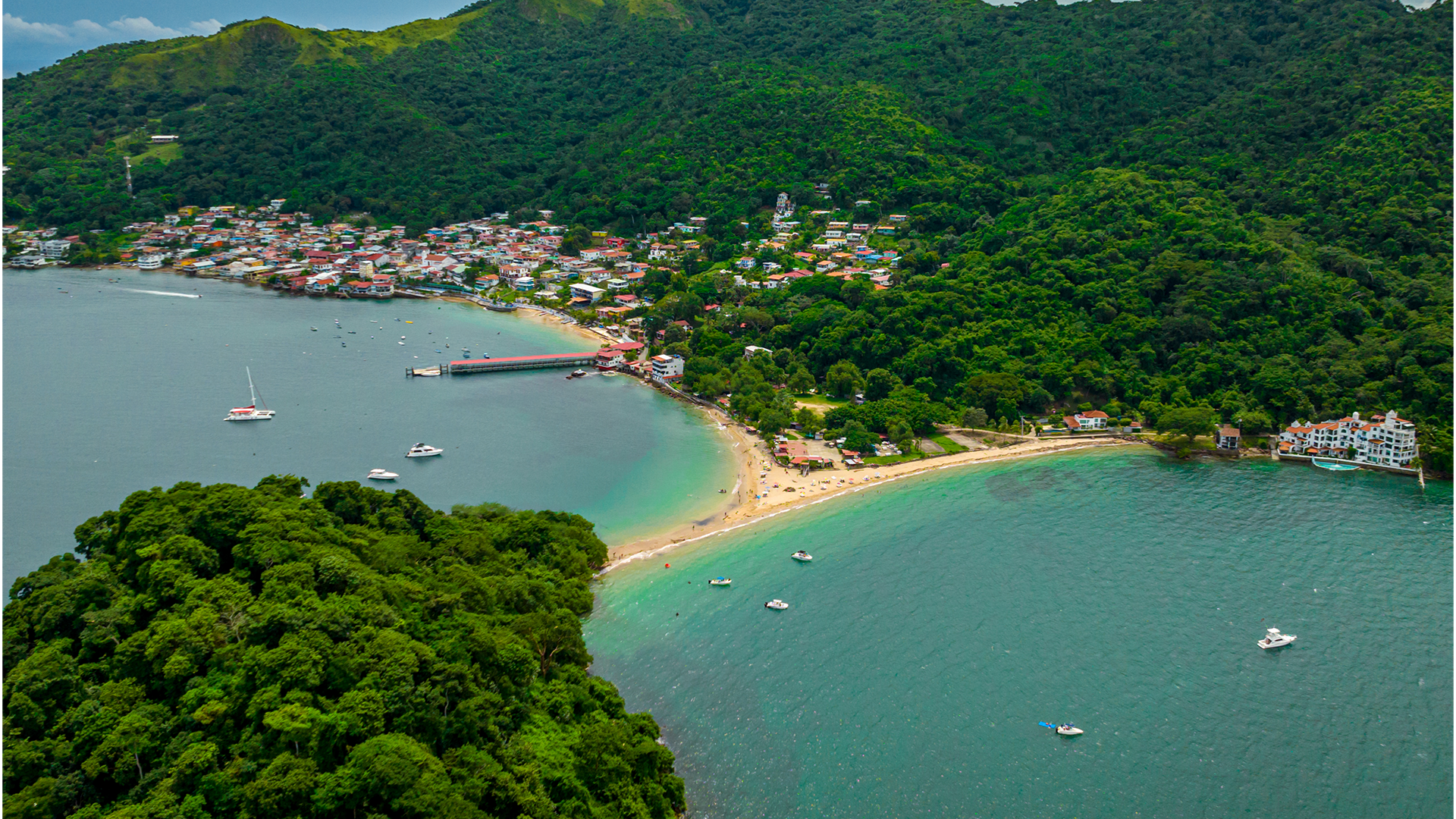 Overhead view of blue ocean and a beach with houses and green forestland further ashore