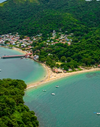 Overhead view of blue ocean and a beach with houses and green forestland further ashore