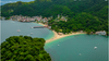 Overhead view of blue ocean and a beach with houses and green forestland further ashore