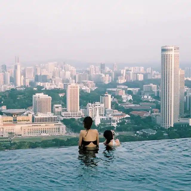Two female swimmers seen from behind at the edge of the infinity pool on the rooftop of the Marina Bay Sands hotel, Singapore, overlooking the city skyscrapers below