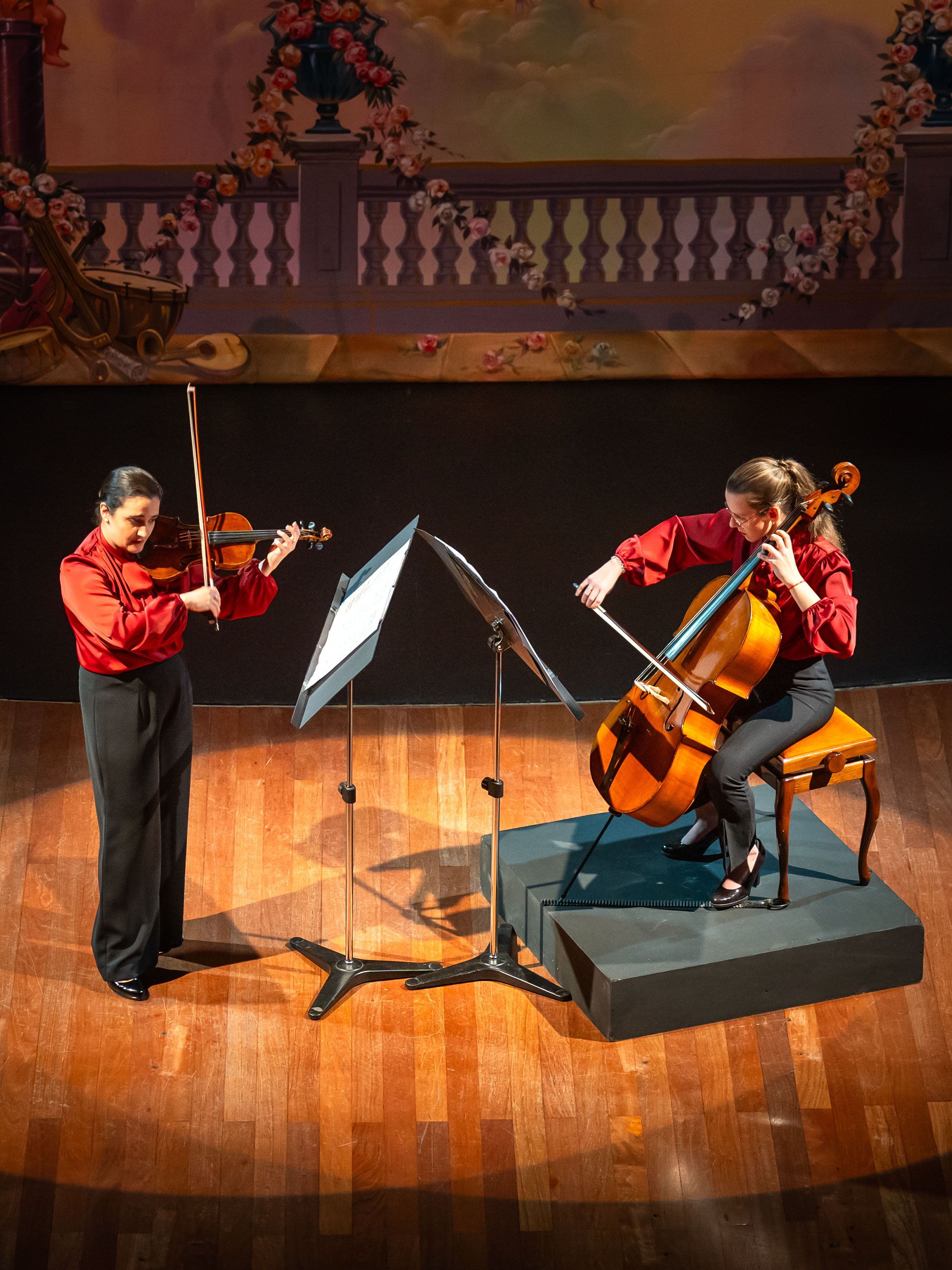 Two musicians perform violin and cello on a theater stage with painted backdrop.